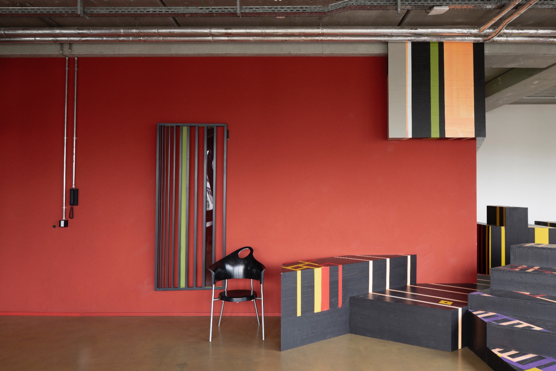 Deep red wall with a black striped panel, a black chair and a suspended colour block